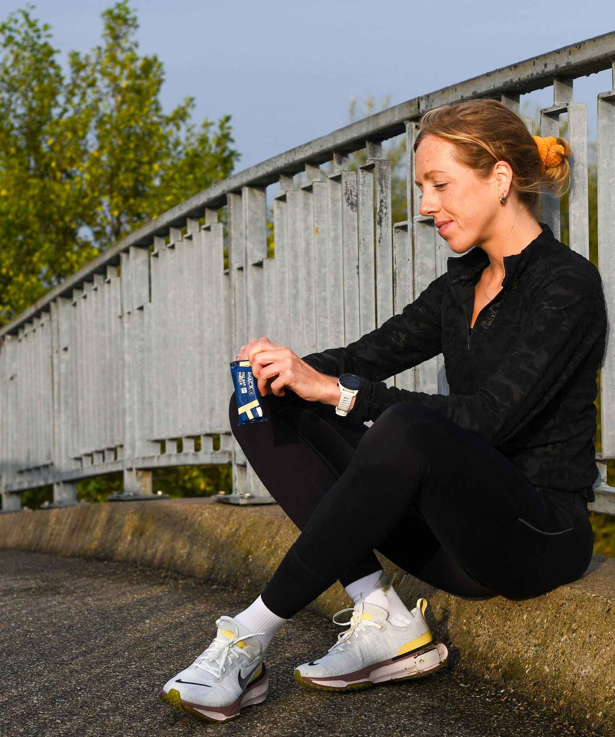 Woman sitting outdoors opening Amacx Fast Bar Vanilla snack after running in athletic wear on a bridge path