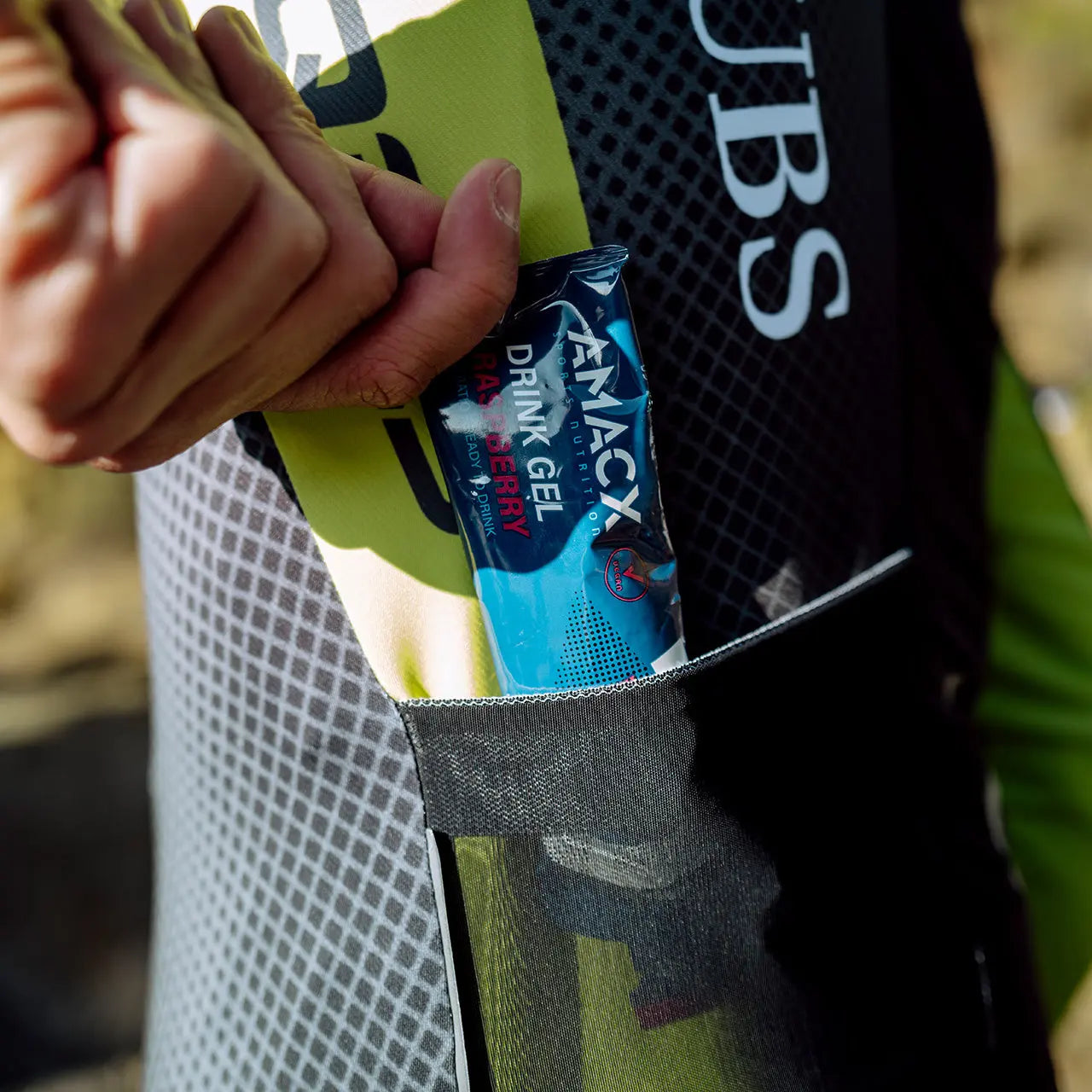 Hand placing Amacx Drink Gel Raspberry into a cycling jersey pocket during outdoor activity.
