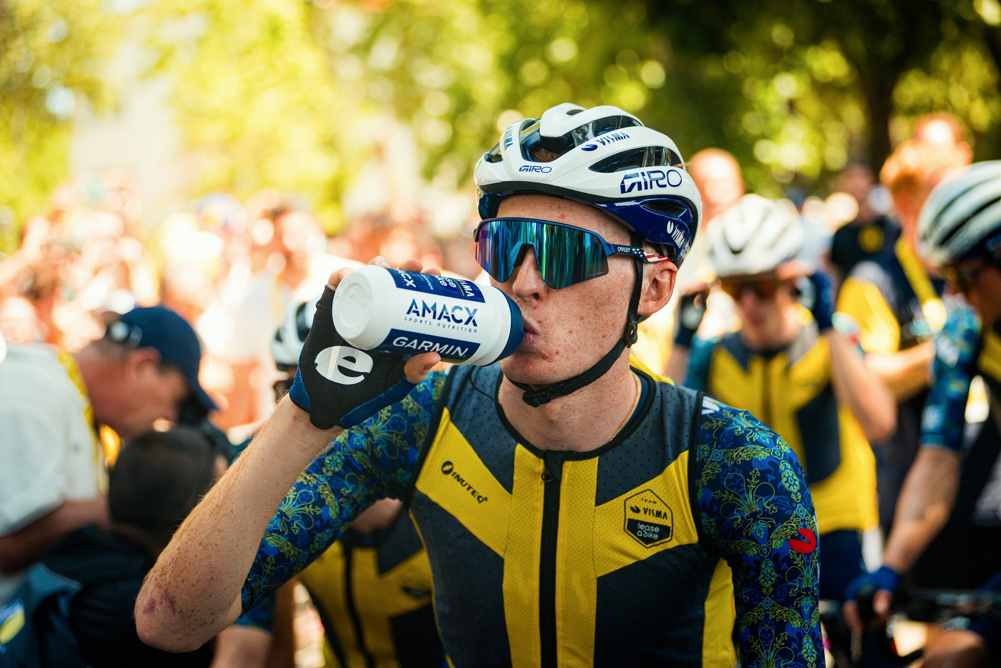 Cyclist wearing helmet and sunglasses drinks from a bottle labeled Amacx Energy Drink | 1.0 kg during event outdoors