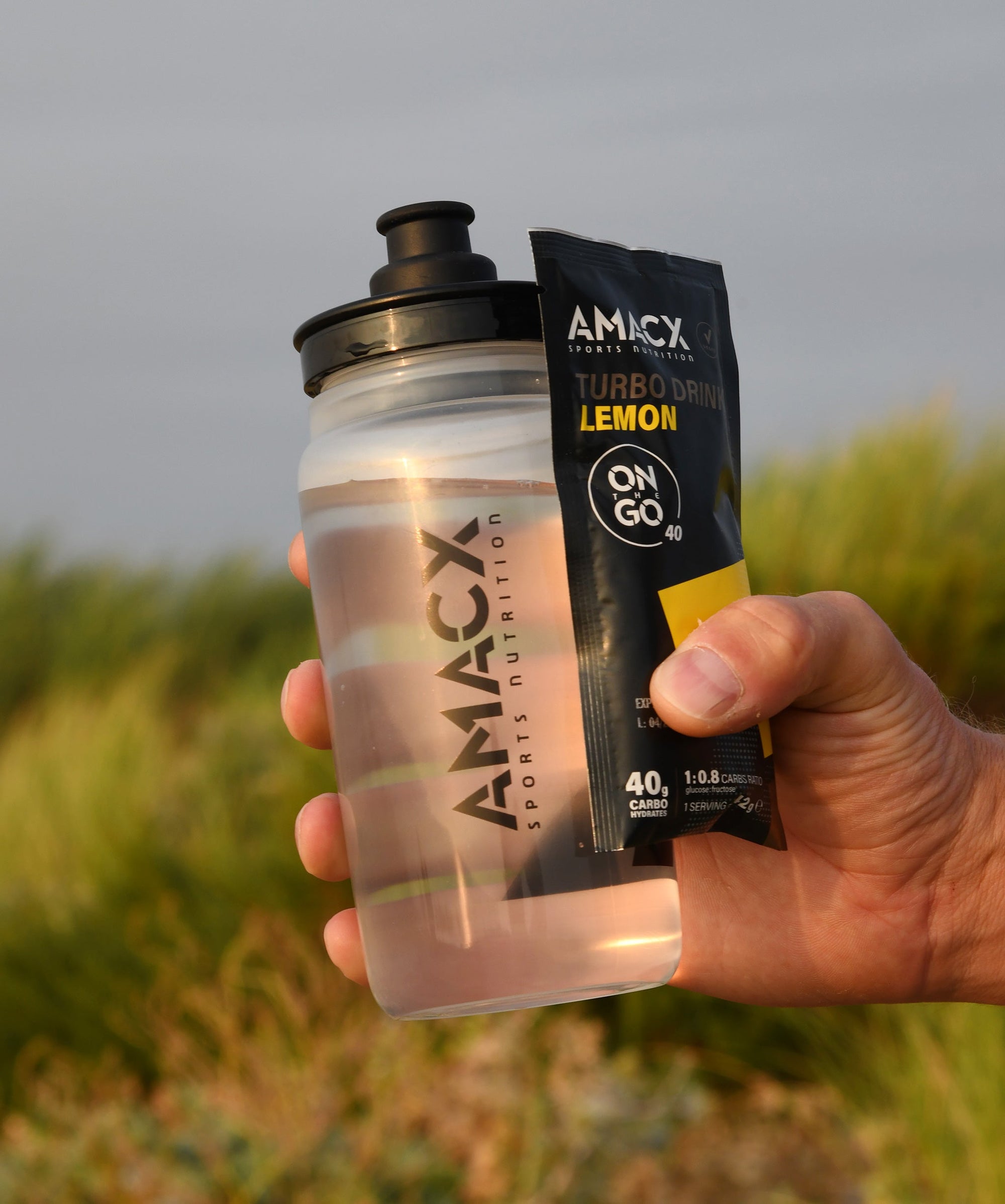 Hand holding Amacx Turbo Drink On The Go Lemon packet and a transparent AMACX sports nutrition bottle outdoors