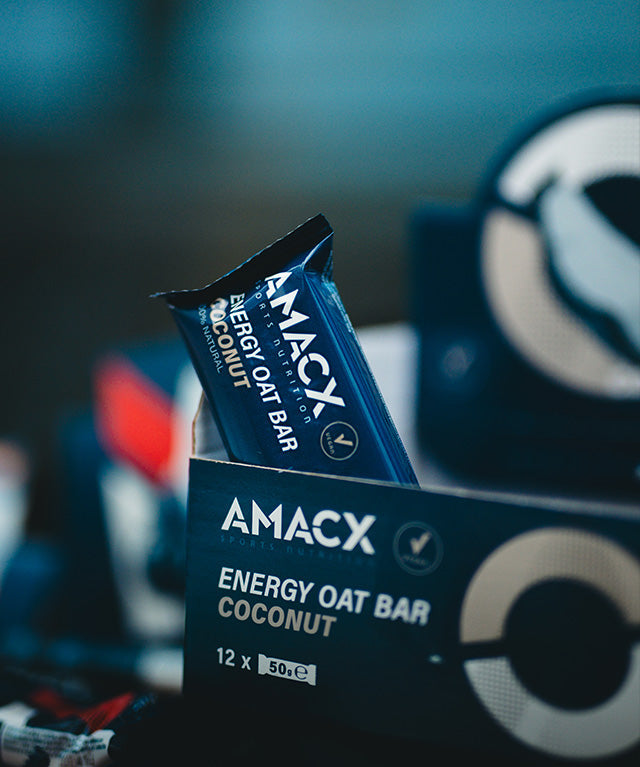 Amacx Energy Oat Bar Coconut packed in a dark blue wrapper partially inside a branded box with blurred background.