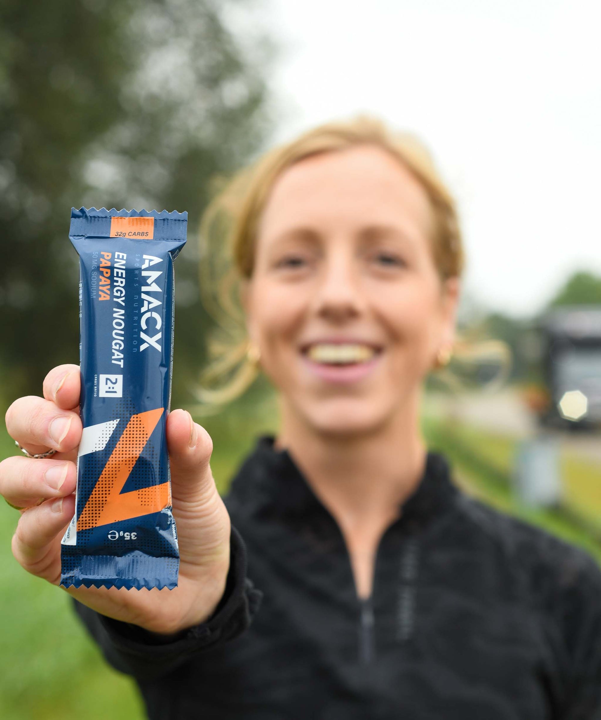 Woman holding Amacx Energy Nougat Papaya energy bar outdoors with blurred background and smiling face