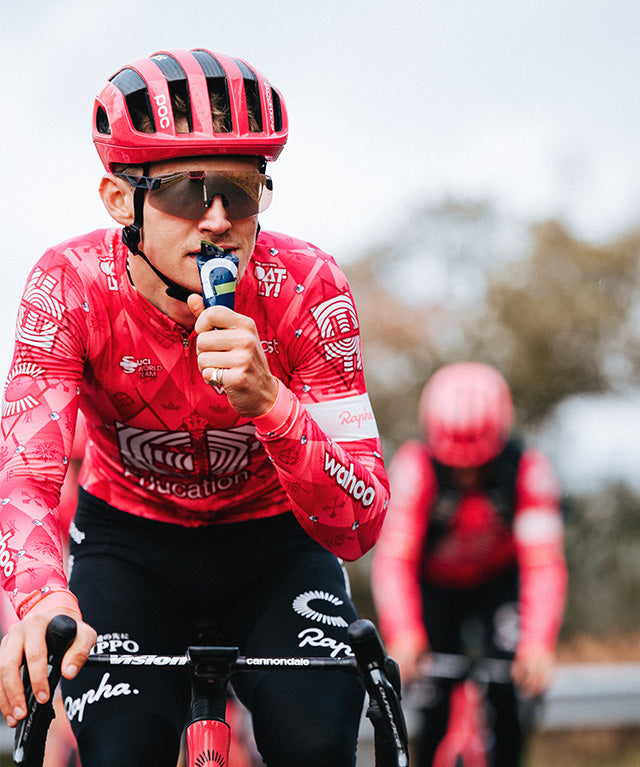 Cyclist wearing red gear consumes Amacx Drink Gel Citrus while riding a bike outdoors with another cyclist behind.