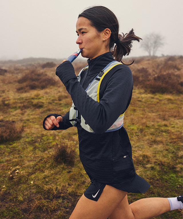 Woman running outdoors drinking Amacx Drink Gel Strawberry from a portable pouch during exercise