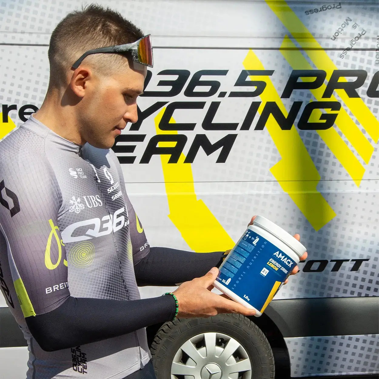 Athlete in cycling gear holding a container of Amacx Energy Drink | 1.0 kg by a cycling team vehicle.