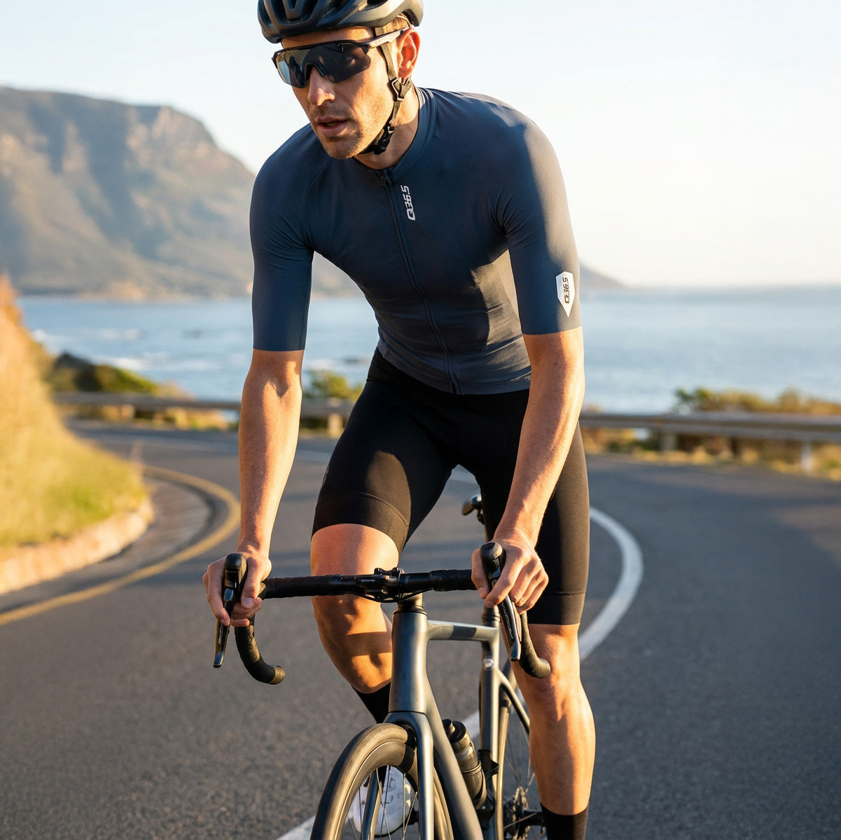 Man cycling on road wearing Q36.5 Gregrarius Pro Jersey - Men's - China Blue with scenic coastal background