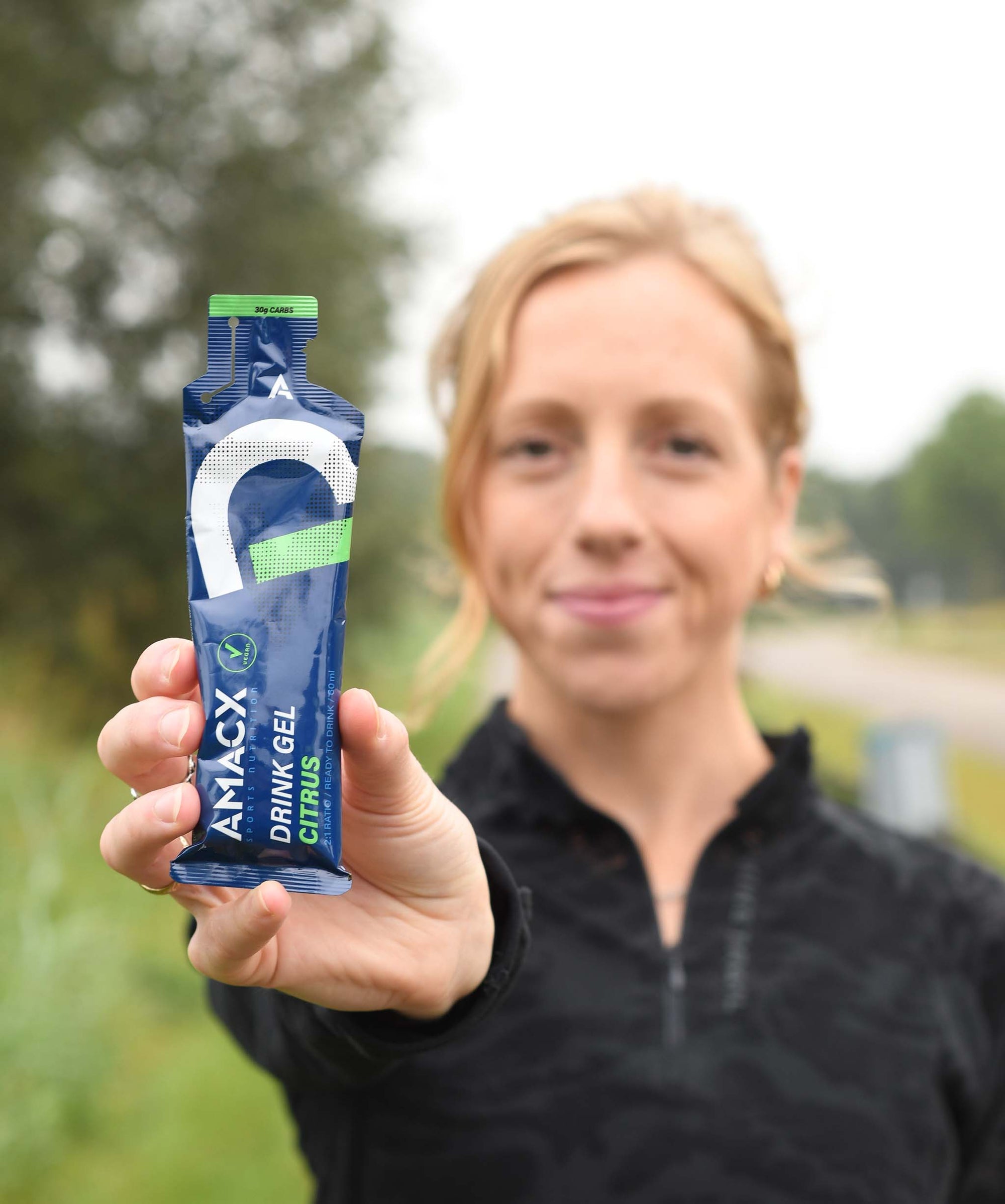 Woman holding a blue packet of Amacx Drink Gel Citrus outdoors, likely after or during exercise.