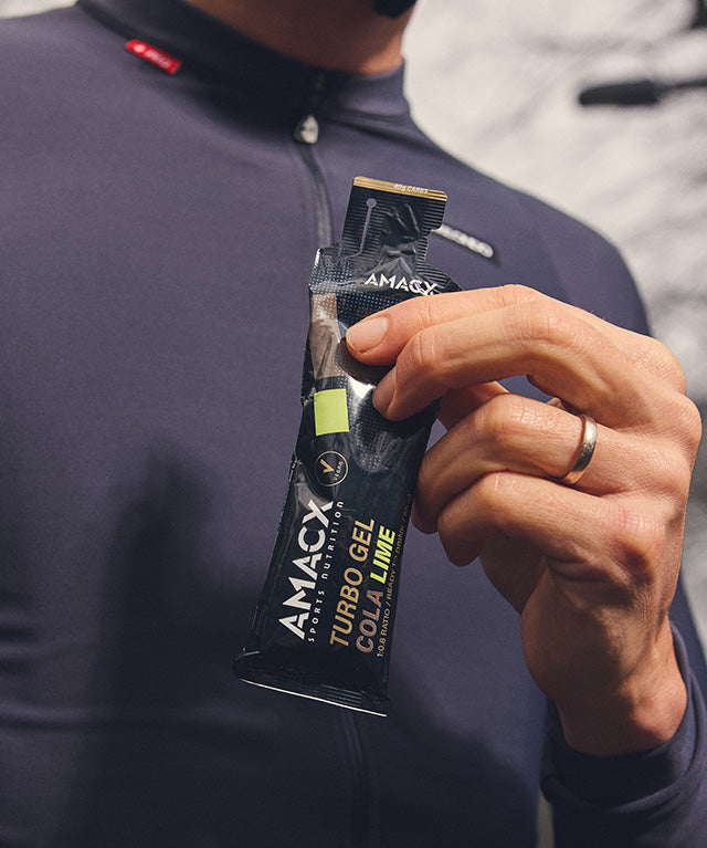 Person holding a black packet of Amacx Turbo Gel Cola Lime energy gel with a dark athletic top background