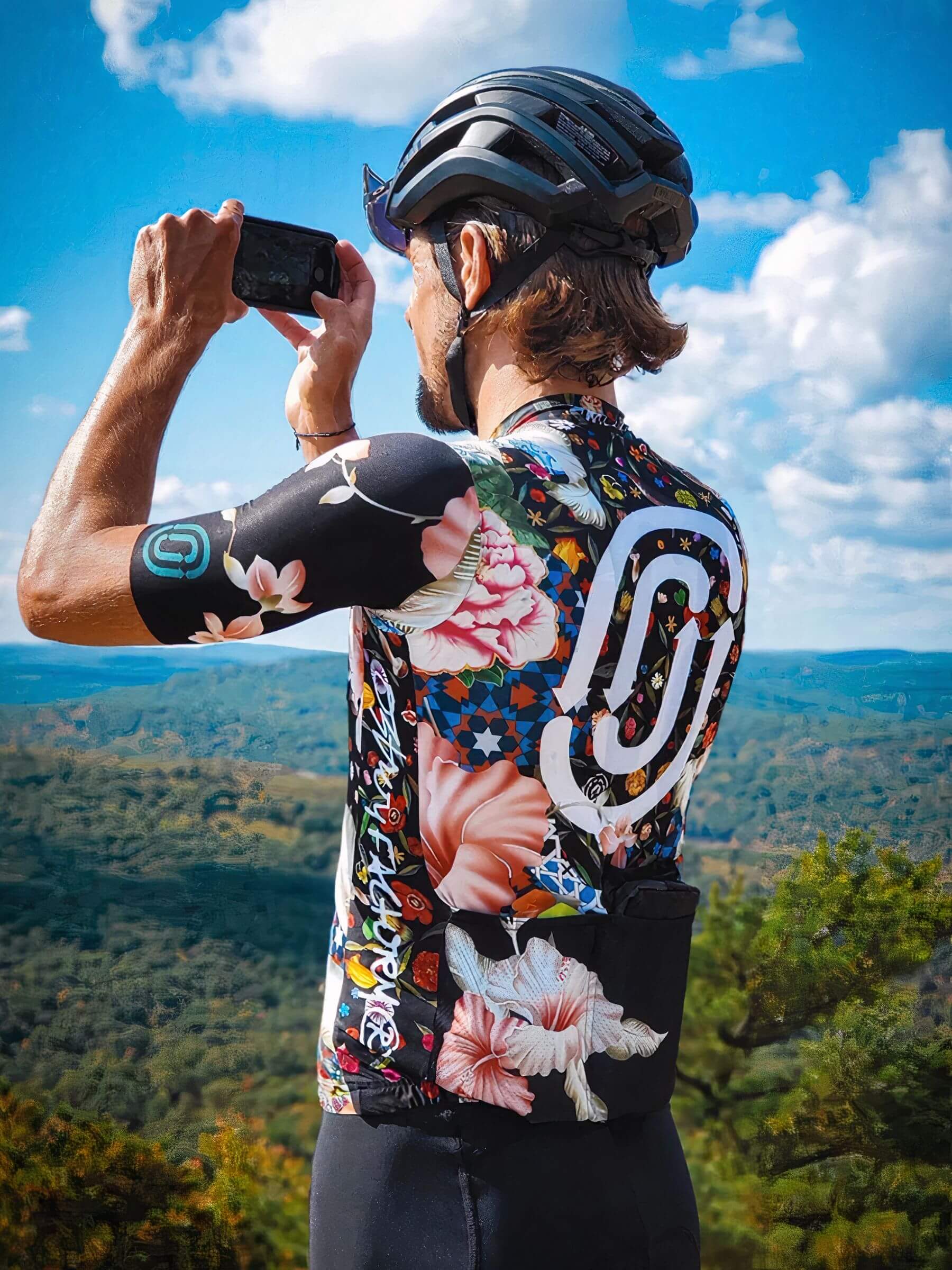 Man wearing Ostroy Omloop Jersey - Men's with floral design taking photo outdoors against scenic landscape