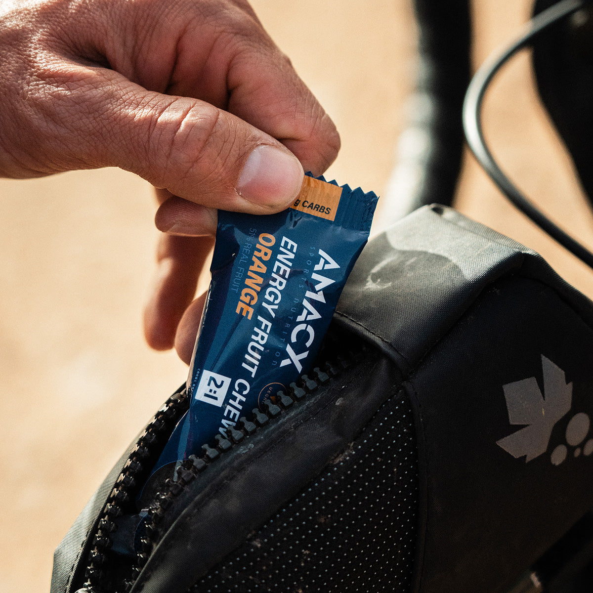 Hand placing Amacx Energy Fruit Chew Orange packet into a black zippered bag during outdoor activity