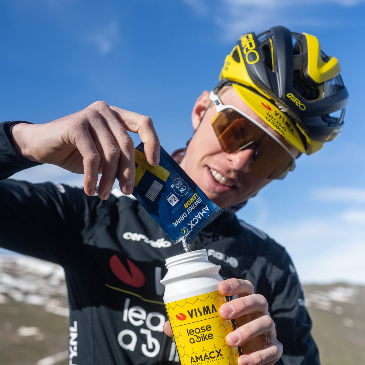 Cyclist wearing helmet and sunglasses pouring Amacx Energy Drink On The Go into a water bottle outdoors on a sunny day