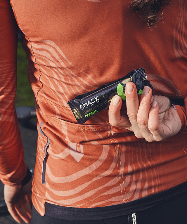 Person wearing orange athletic wear placing Amacx Turbo Fruit Chew Citrus in their back pocket during outdoor activity