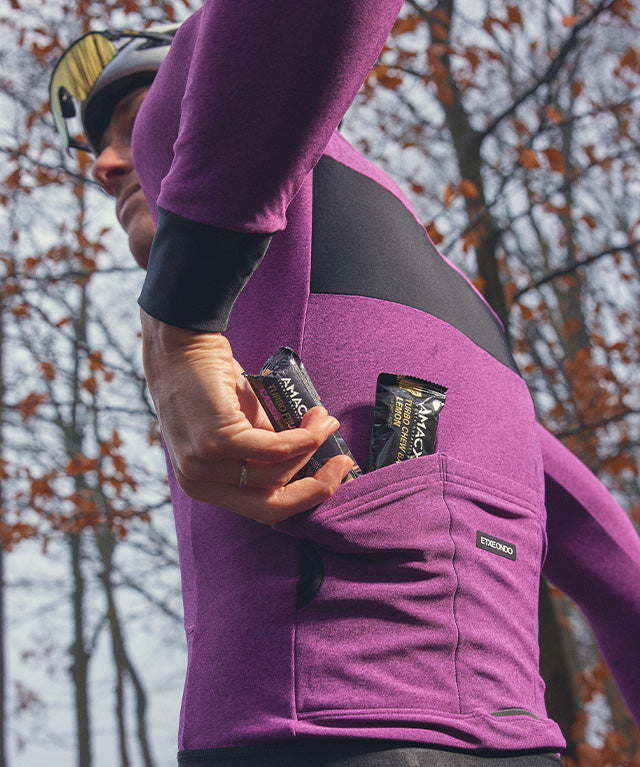 Person wearing purple cycling jersey placing Amacx Turbo Fruit Chew Cassis bars into back pocket outdoors
