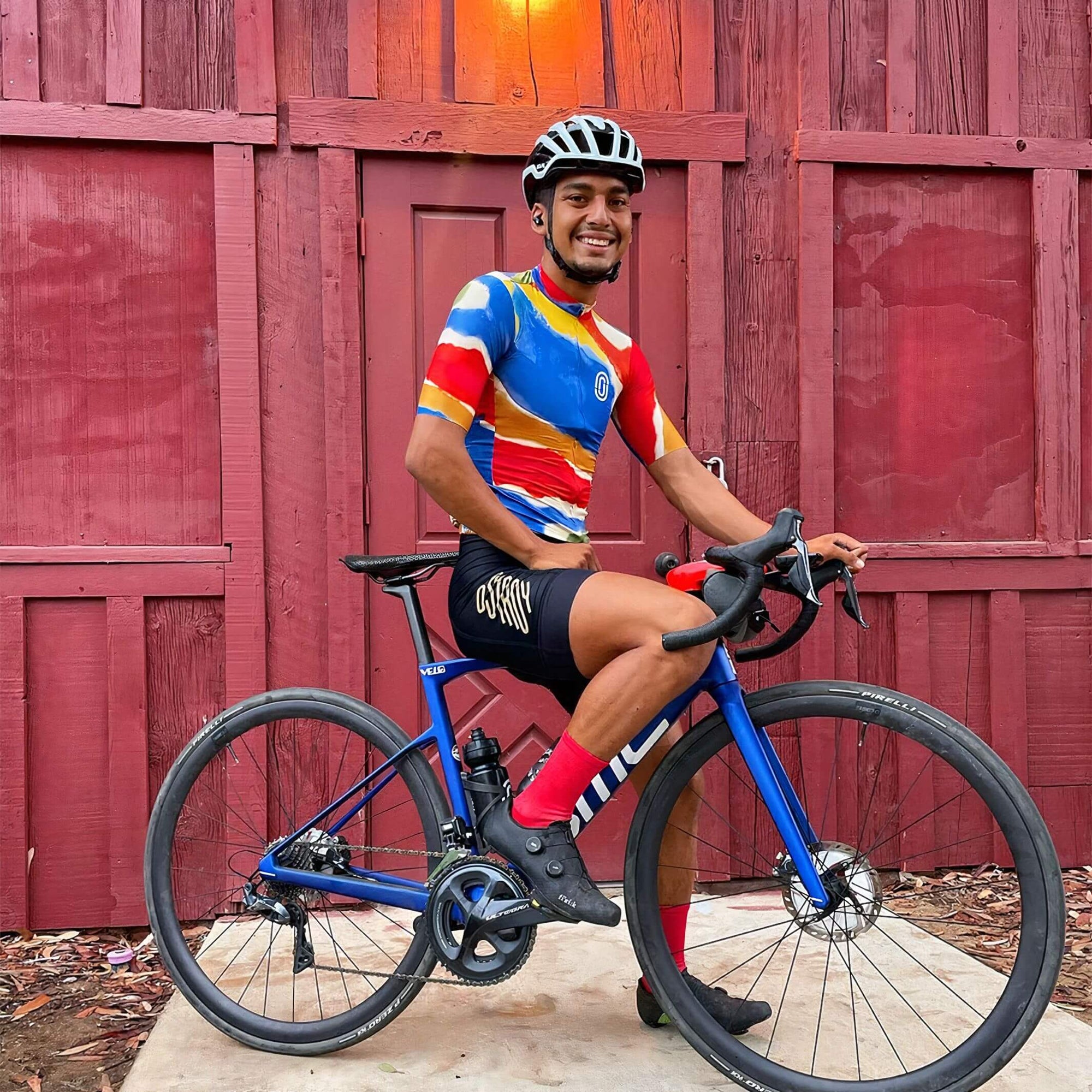 Man wearing Ostroy Watercolor Jersey - Men's with vibrant abstract design sitting on blue road bike against red wooden wall